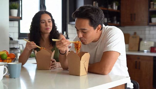 Couple enjoying takeout noodles at kitchen counter using chopsticks casual home dining