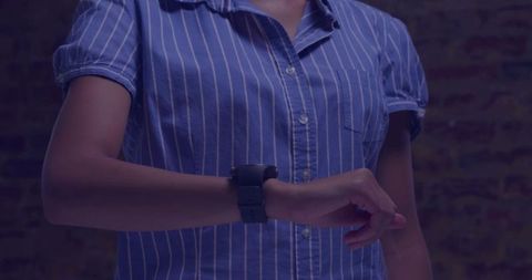 Woman adjusting smartwatch on left wrist wearing blue striped shirt under moody lighting