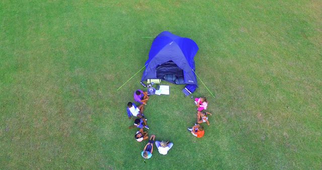 Top view of kids sitting in a circle near a tent on a grassy field. Perfect for articles about outdoor education, group activities, and children's summer camps. Ideal for illustrating teamwork, adventure, and bonding in nature settings.