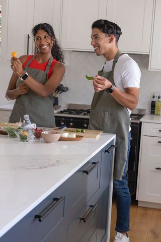 African couple cooking at modern kitchen island wearing green aprons, chopping peppers