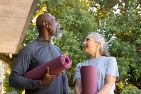 Mature black man and white woman holding yoga mats looking upward on sunlit porch