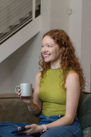 Smiling redhead woman sipping coffee on sofa holding smartphone, casual home lifestyle