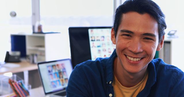 Young man smiling in modern office environment with computer screens in background. Ideal for workplace culture articles, professional success stories, or advertisements highlighting modern work environments.
