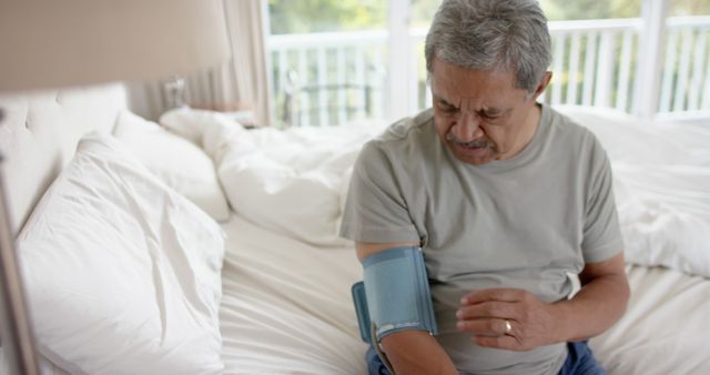 Elderly man sits on bed wearing casual clothes, monitoring blood pressure with digital device. Looks serious about health. Useful for healthcare, senior wellness, home health monitoring themes.