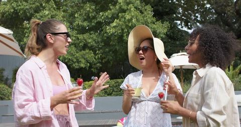 Diverse women chatting and enjoying poolside cocktails, wearing sun hats and shades