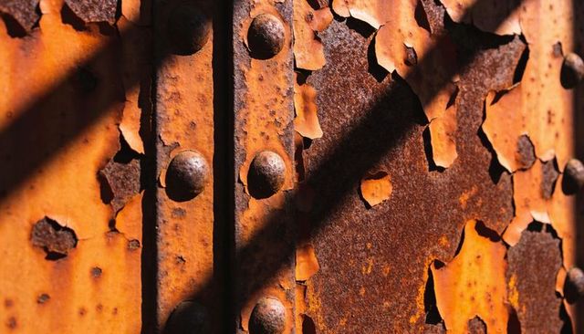 Peeling orange paint revealing rusted riveted steel girder with diagonal shadow