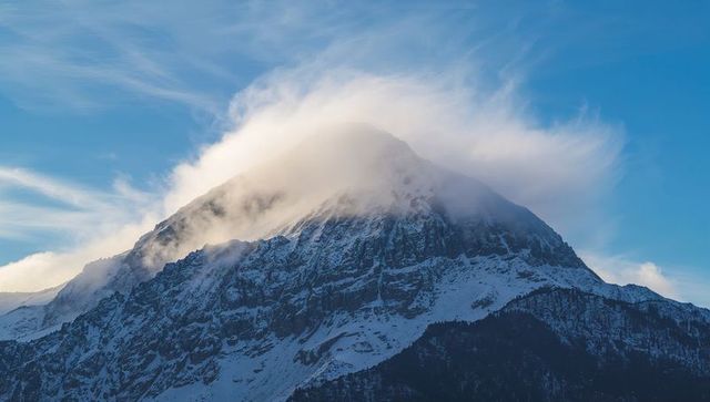 Snowcapped mountain peak rising with dramatic cloud cap over alpine ridge at dawn