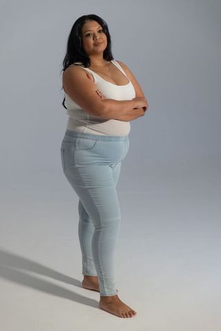 Asian plus-size woman standing barefoot in white tank and light blue jeans, arms crossed, confident