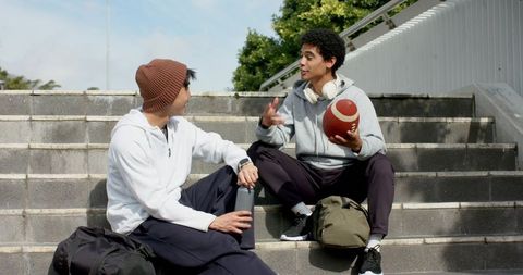 Asian and african american friends chatting on campus steps holding football and bottle
