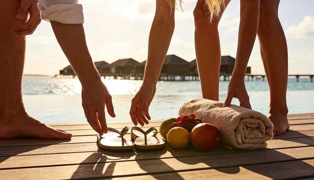 Couple reaching for sandals on tropical dock at golden hour with towel and fruit