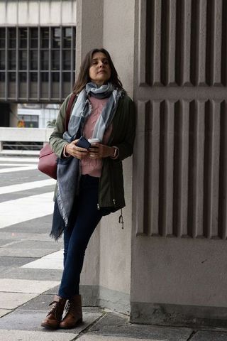 Middle-aged woman leaning on column at urban crosswalk, waiting with coffee and smartphone