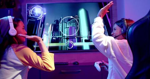 Two teenage girls are sitting in front of a colorful screen, actively playing a video game while wearing headsets. They appear engaged and are sharing a moment of excitement, exemplifying the use of technology for entertainment and social interaction. This image is suitable for illustrating themes of youth culture, technological bonding, online gaming communities, and friendship. Perfect for advertising gaming products, headsets, entertainment systems, and youth engagement in technology.