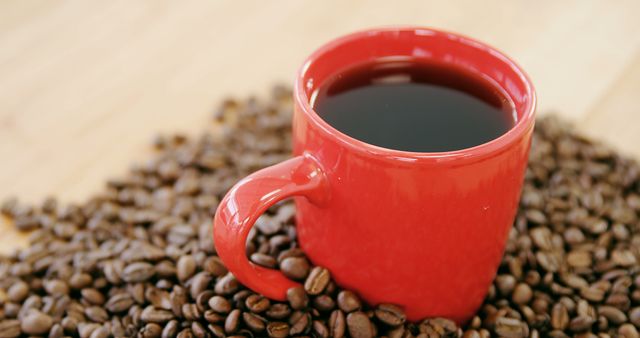 A red mug filled with coffee sits amidst a scattering of coffee beans on a wooden surface. The warm tones and the inviting cup of coffee suggest a cozy, energizing break or the start of a morning routine.