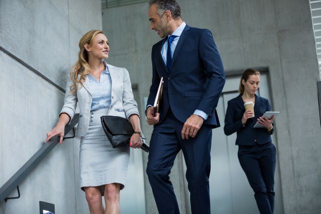 Business professionals engaging in conversation while descending stairs in a modern office building. Ideal for illustrating corporate communication, teamwork, and professional environments. Useful for business-related articles, corporate websites, and promotional materials.