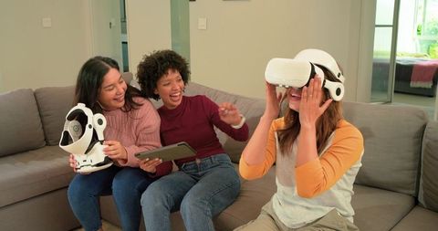 Diverse women trying virtual reality headset on sofa laughing and gaming together