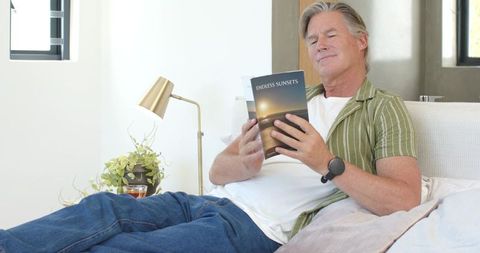 Mature man reclining on couch reading book and relaxing at home in sunlit living room
