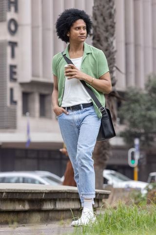 African american man walking on low ledge with coffee cup and crossbody bag in urban plaza