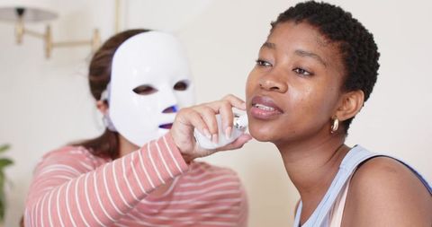 African woman receiving at-home led facial treatment by therapist wearing light mask