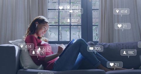 Relaxing woman reclining on sofa using tablet with stylus and floating social notifications