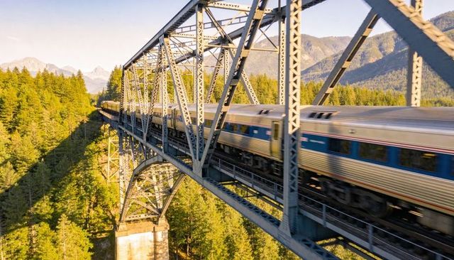 Crossing Passenger Train Spanning Steel Truss Bridge Above Evergreen Forest and Mountains