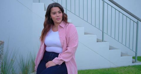 Young woman sitting by outdoor stairs wearing pastel pink shirt and high-waisted jeans