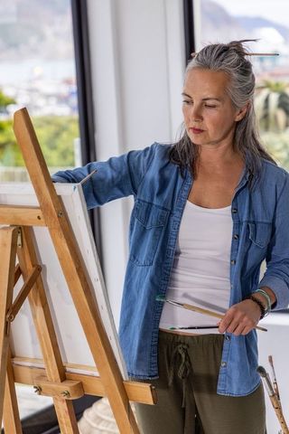 Mature woman painting on canvas in sunlit coastal studio overlooking marina harbor