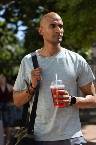 Man standing in sunlit urban plaza wearing gray t-shirt holding red iced drink and shoulder bag