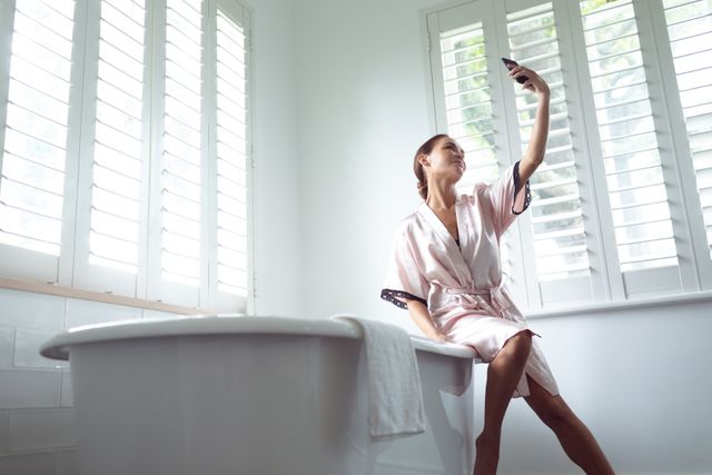 Woman sitting on edge of bathtub taking selfie in bright, modern bathroom. Ideal for lifestyle, self-care, and home decor concepts. Perfect for promoting beauty, relaxation, and modern living.