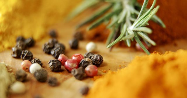 Various peppercorns including red, black, and white are scattered on a wooden surface alongside sprigs of fresh rosemary. The vibrant colors of the peppercorns contrast with the green of the rosemary, creating an appealing display for culinary contexts. Ideal for use in articles, blogs, recipe books, and websites promoting cooking, healthy eating, and natural food ingredients.