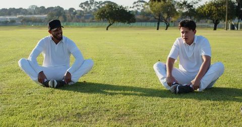 Diverse teammates doing butterfly stretch on grass field outdoor cricket training in warm sunlight