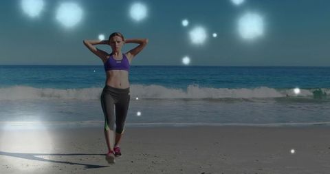Woman walking on sandy beach with hands behind head exercising in sports bra and leggings