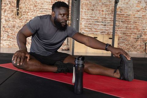 African american man stretching leg on red mat in industrial gym with shaker bottle
