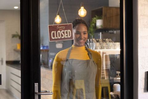 African american barista smiling behind cafe door with closed sign and warm cozy interior