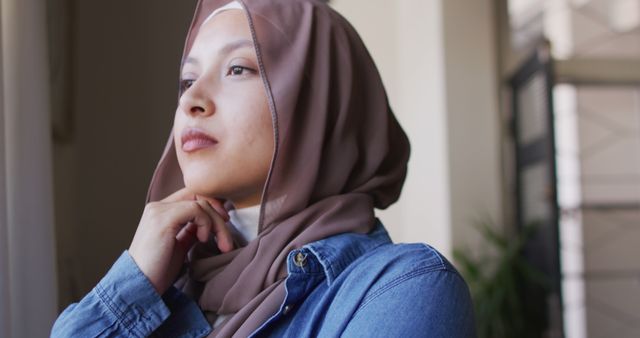 Image of thoughtful biracial woman in hijab looking out of window at home. Relaxation, inclusivity and domestic life.
