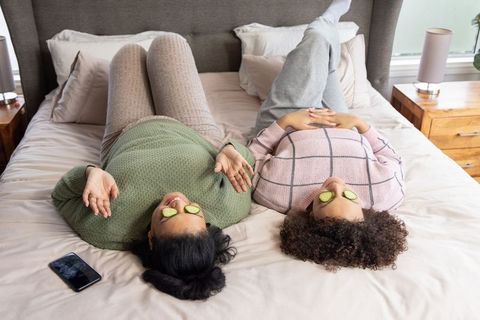 Multiracial friends relaxing with cucumber eye masks on bed enjoying cozy self-care moment