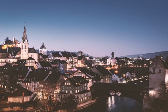 Image captures a picturesque evening scene of a historic European town with well-lit buildings and a church by the riverside. Village streets and houses are perfectly aligned, illuminated against a backend dusk sky, providing a serene and charming nightscape. Ideal for travel websites, postcards, blog posts about European architecture or history, or inspirational city skyline posters.