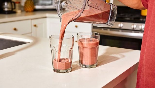 Pouring pink fruit smoothie from blender into two glasses on modern white kitchen counter