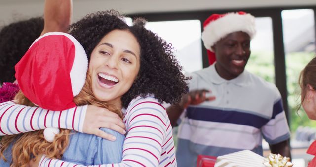 Friends embrace in warm, joyful moment while celebrating Christmas; people wearing Santa hats and exchanging gifts with smiles. Perfect for holiday greeting cards, festive advertisements, and multicultural holiday campaigns emphasizing joy and togetherness.