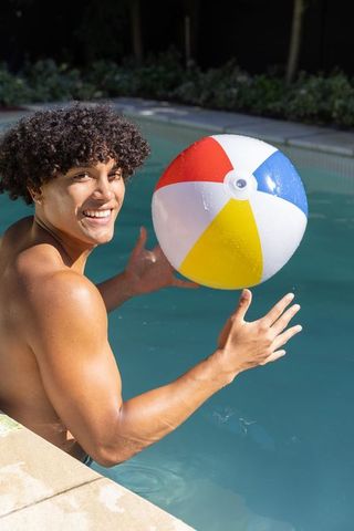 Shirtless young man smiling and playing with colorful beach ball in backyard pool