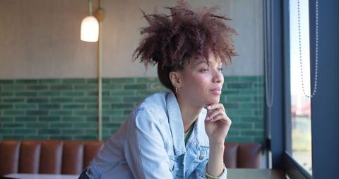 Young woman gazing out cafe window leaning on hand wearing denim jacket reflecting quietly