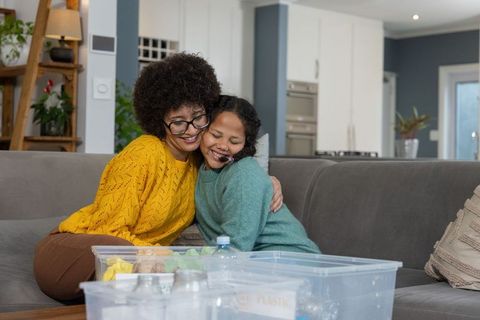 Mother and daughter cuddling on gray sofa sharing hug and sorting recycling bins at home