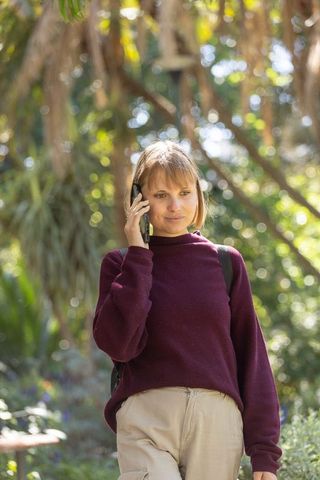 Young woman talking on smartphone in leafy park wearing maroon sweater and backpack