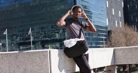 African American Man Leaning on City Railing Adjusting Headphones Carrying Duffel Bag
