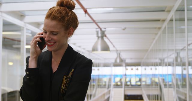 Red-haired businesswoman dressed in professional attire is seen happily talking on her phone while standing in a sleek and modern office corridor. Could be used for business, telecommunications, corporate professionalism, and workplace diversity content.