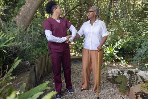 Caregiver supporting african american senior walking with cane on garden path