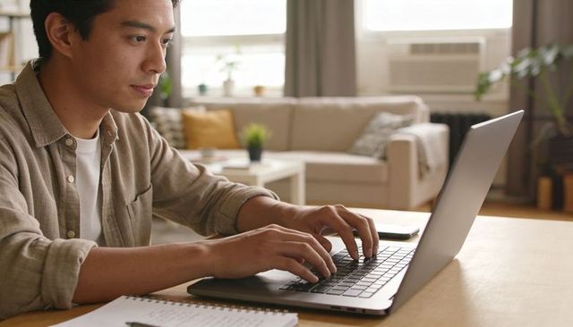 Young man typing on laptop in cozy sunlit home workspace, focused remote work lifestyle