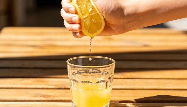 Hand squeezing lemon half over glass on wooden table in warm sunlight juice droplets