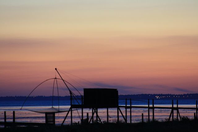 Silhouetted fishing nets on pier during sunset, creating tranquil and serene scene. Ideal for nature or travel articles, peaceful mood photography, evening landscape prints, or backgrounds depicting calm and relaxing environments.
