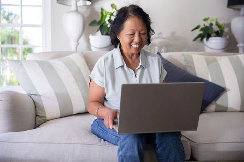 Senior asian woman smiling while using laptop on cozy striped sofa in bright living room