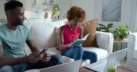 African american couple on sofa, woman writing in notebook, man using phone and laptop